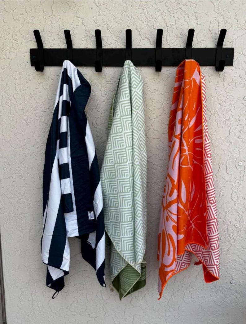K&ouml;nnte beinhalten: Eine schwarze Wandgarderobe mit sieben Haken h&auml;lt drei h&auml;ngende Handt&uuml;cher. Das linke Handtuch hat marineblaue und wei&szlig;e vertikale Streifen. Das mittlere Handtuch zeigt ein hellgr&uuml;nes und wei&szlig;es geometrisches Muster. Das rechte Handtuch hat einen orangefarbenen und hellrosa tropischen Blattdruck.