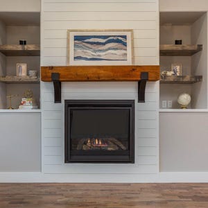 May include: A fireplace with a black frame and a glowing fire. Above the fireplace is a wooden mantel supported by black brackets. A framed landscape painting hangs above the mantel. Built-in shelves are on either side.