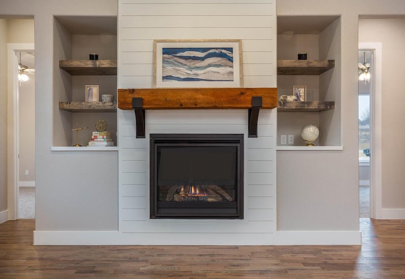May include: A fireplace with a black frame and a glowing fire. Above the fireplace is a wooden mantel supported by black brackets. A framed landscape painting hangs above the mantel. Built-in shelves are on either side.