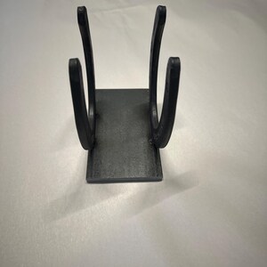 May include: A black metal stand constructed from two vertical horseshoes welded onto a rectangular base plate. The object features a matte finish and a symmetrical design, with the curved ends of the horseshoes facing upward to create a holder.