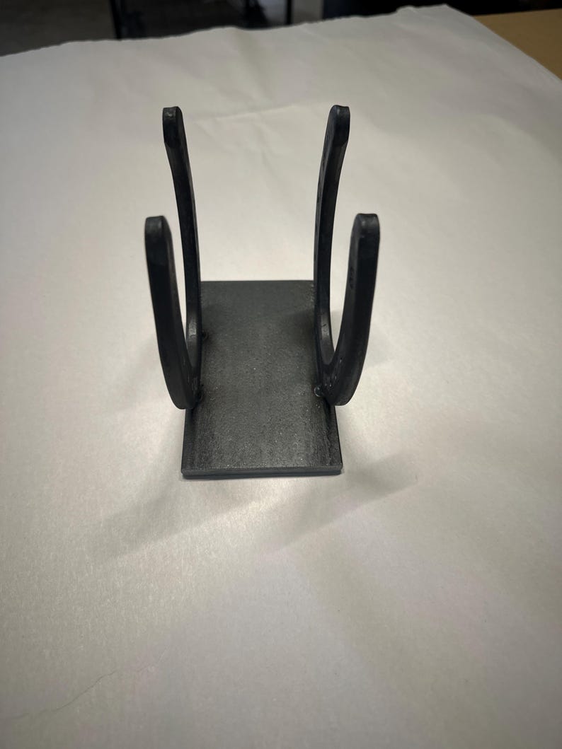 May include: A black metal stand constructed from two vertical horseshoes welded onto a rectangular base plate. The object features a matte finish and a symmetrical design, with the curved ends of the horseshoes facing upward to create a holder.