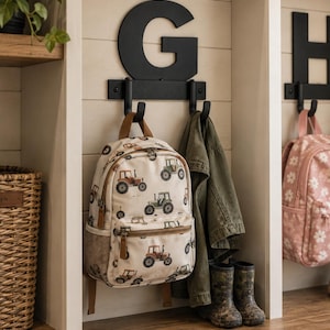 May include: A cream-colored backpack featuring a repeating pattern of small tractors in shades of red and green. The backpack has a front zippered pocket, side mesh pockets, and brown straps. It hangs from a black metal hook mounted below a large letter G.