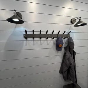 K&ouml;nnte beinhalten: Eine mattschwarze Wandgarderobe mit zehn Haken, die zwischen zwei passenden schwarzen, kegelf&ouml;rmigen Wandleuchten angebracht ist. Eine dunkelblaue Baseballkappe mit einem runden Aufn&auml;her h&auml;ngt an einem Haken, eine dunkelgraue Jacke an einem anderen.