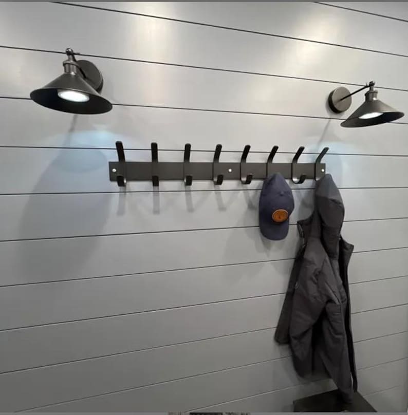 K&ouml;nnte beinhalten: Eine mattschwarze Wandgarderobe mit zehn Haken, die zwischen zwei passenden schwarzen, kegelf&ouml;rmigen Wandleuchten angebracht ist. Eine dunkelblaue Baseballkappe mit einem runden Aufn&auml;her h&auml;ngt an einem Haken, eine dunkelgraue Jacke an einem anderen.