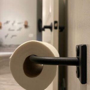 May include: Black metal toilet paper holder mounted on a white door with a roll of white toilet paper.