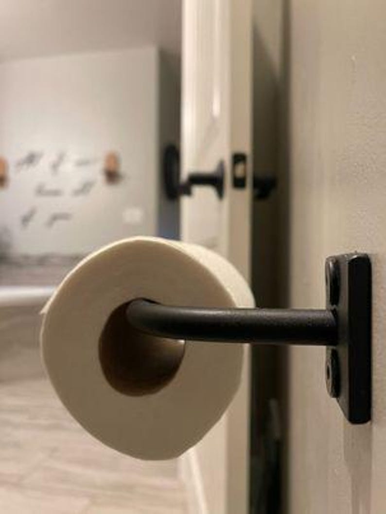 May include: Black metal toilet paper holder mounted on a white door with a roll of white toilet paper.