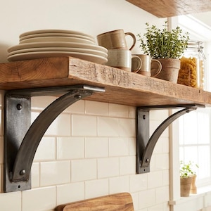 May include: A thick, natural-toned wooden shelf supported by two dark, curved metal brackets. The shelf holds a stack of cream-coloured plates, three ceramic mugs in earthy tones, a small potted green plant, and a glass jar filled with pasta.