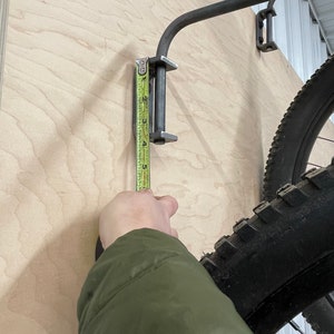 May include: A metal bike rack mounted to a wall with a measuring tape showing the height of the rack is 4 inches. The rack is designed to hold a bicycle tire.