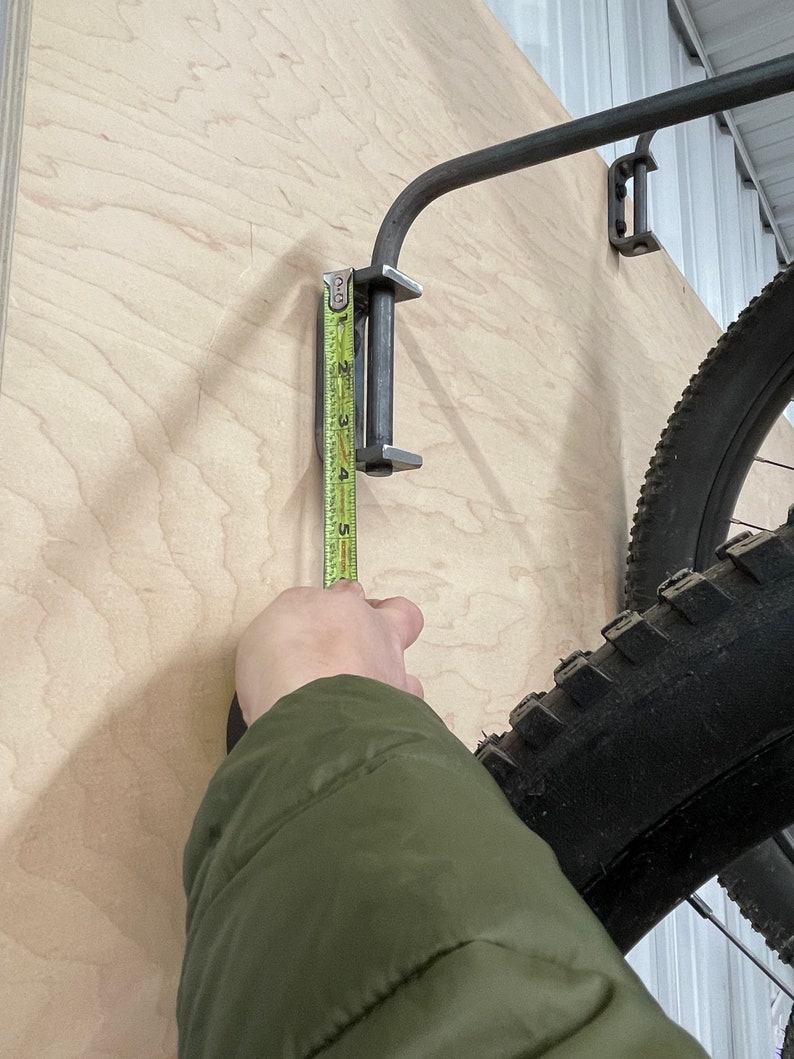 May include: A metal bike rack mounted to a wall with a measuring tape showing the height of the rack is 4 inches. The rack is designed to hold a bicycle tire.