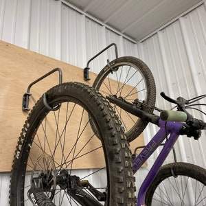 May include: Two bicycles are hanging on a wall mounted bike rack. The bikes are both black with purple frames. The bike on the left has a black tire with white lettering that says "Maxxis" and the bike on the right has a black tire with white lettering that says "Prevail".
