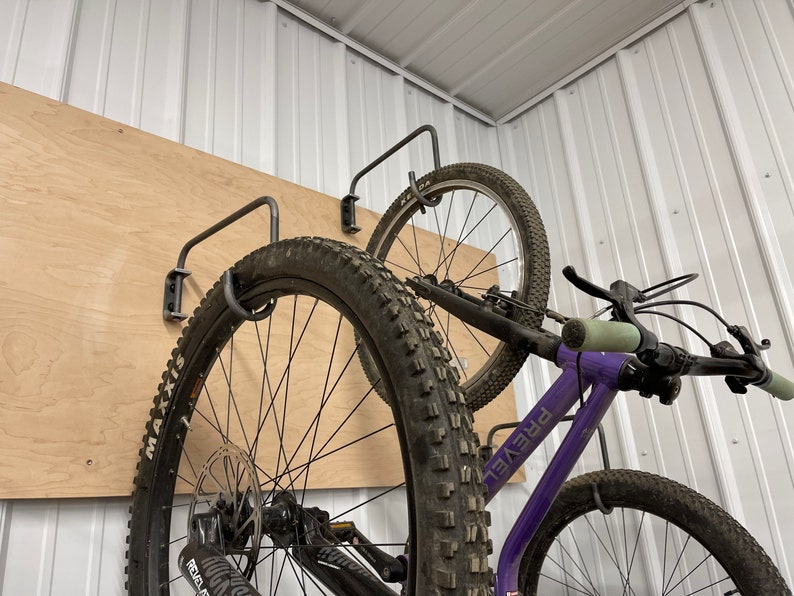 May include: Two bicycles are hanging on a wall mounted bike rack. The bikes are both black with purple frames. The bike on the left has a black tire with white lettering that says "Maxxis" and the bike on the right has a black tire with white lettering that says "Prevail".