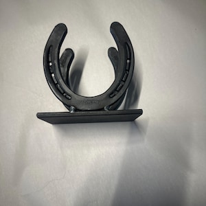 May include: A matte black metal holder constructed from two U-shaped horseshoes welded together in a vertical orientation, attached to a flat rectangular base plate. The design features visible nail holes along the curved edges of the horseshoes.