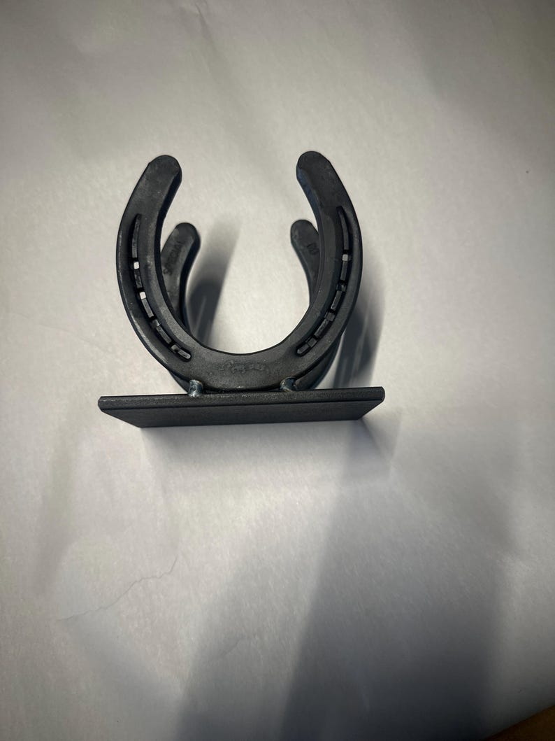 May include: A matte black metal holder constructed from two U-shaped horseshoes welded together in a vertical orientation, attached to a flat rectangular base plate. The design features visible nail holes along the curved edges of the horseshoes.