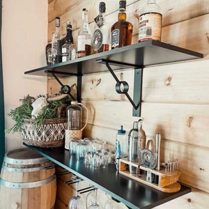 May include: Black floating shelves with metal brackets, holding various bottles of liquor, glasses, and bar tools. The shelves are mounted on a wood wall.