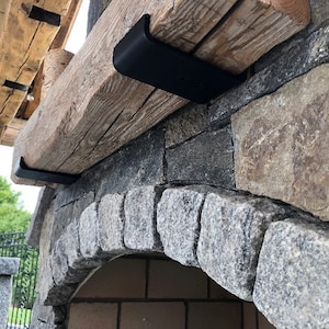 May include: A heavy-duty matte black metal bracket supporting a thick, weathered wooden beam mantel. The bracket is mounted against a textured stone fireplace surround, featuring a curved archway constructed from individual gray stone blocks.