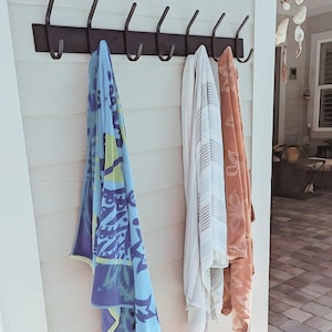 May include: A black metal rack with six hooks mounted on a white wall. Three towels are hanging on the hooks, one blue with yellow and white patterns, one white with gray stripes, and one orange with a floral pattern.