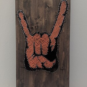 Peut inclure: Une œuvre d'art en fil sur une planche de bois représentant une main faisant le geste du rock and roll. La main est faite de fil orange et le fond est noir.