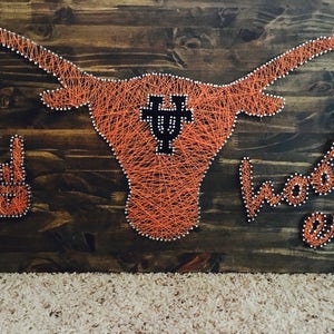 May include: Orange string art on a wooden background depicting a longhorn steer with the University of Texas logo in the center. The words "Hook 'em" are also spelled out in string art.