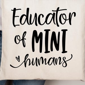 May include: Cream-colored tote bag with the text "Educator of MINI humans" in black script. The bag is made of a sturdy fabric and has a simple design, suitable for carrying books or supplies.