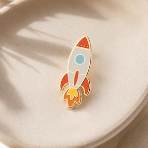 May include: A gold-toned enamel pin shaped like a rocket ship. The rocket features a light gray body, a red nose cone with small dots, a light blue circular window, red side fins, and a bright yellow and orange flame at the base.