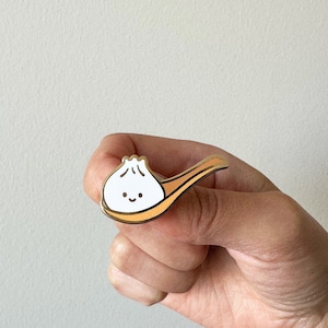 May include: Gold-tone enamel pin featuring a cartoon dumpling with a smiling face, sitting on a gold-tone spoon.