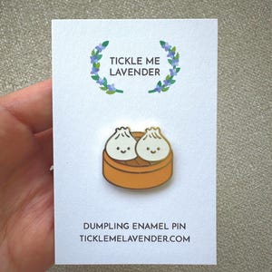May include: An enamel pin featuring two white dumplings with simple black facial features inside a round orange bamboo steamer basket, attached to a white display card with the text TICKLE ME LAVENDER and DUMPLING ENAMEL PIN TICKLEMELAVENDER.COM.