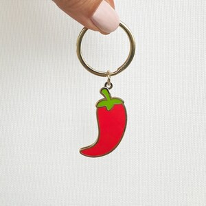 May include: A gold key chain with a red chili pepper charm. The chili pepper is outlined in gold and has a green stem.