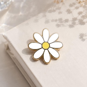 May include: A daisy-shaped enamel pin with a gold-colored outline, white petals, and a yellow center. The pin is resting on a light-colored book. The image also includes a glass of water and dried flowers.