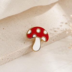 May include: A red and white mushroom enamel pin with a gold-colored outline. The pin features a classic mushroom design with white spots on the red cap and a white stem. The pin is set against a neutral background.