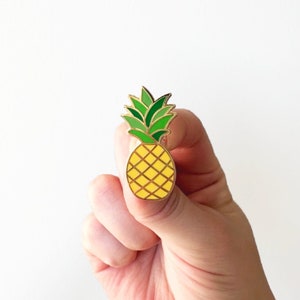 May include: A gold-colored enamel pin featuring a yellow pineapple with green leaves.