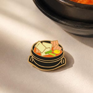 Peut inclure: Une broche en émail noir en forme de bol en terre cuite coréen traditionnel rempli de ragoût de kimchi. La broche présente des cubes de tofu blancs, des tranches de viande roses, un jaune d'œuf jaune vif et des oignons verts dans un bouillon orange.