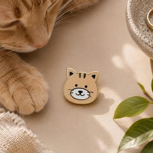 Op de afbeelding: Een goudkleurige emaille speld in de vorm van een kattenkop met een lichtbruin gezicht, donkerbruine strepen op het voorhoofd, zwarte oren en een witte snuit met een zwarte neus en snorharen, geplaatst naast een kattenpoot.