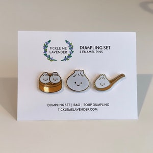 May include: A set of three enamel pins featuring cartoon dumplings. The pins are gold-colored and depict a steamed bun, a dumpling in a steamer basket, and a soup dumpling in a spoon. The text "Dumpling Set" and "3 Enamel Pins" is printed on the white card.