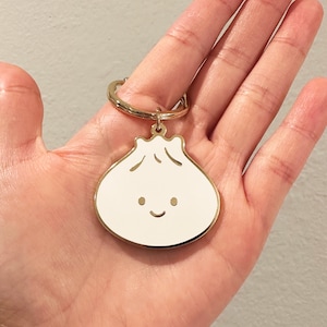 May include: A gold-colored keychain with a white enamel charm shaped like a smiling dumpling.