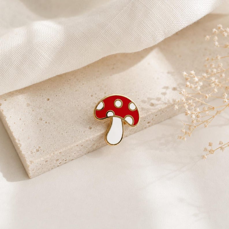May include: A red and white mushroom enamel pin with a gold-colored outline. The pin features a classic mushroom design with white spots on the red cap and a white stem. The pin is set against a neutral background.