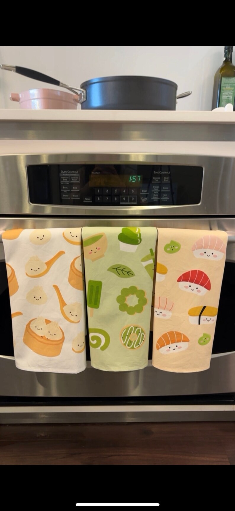 May include: Three kitchen towels hanging on an oven door handle. The towels are white with different designs. The first towel has a pattern of steamed buns with happy faces. The second towel has a pattern of green tea desserts with happy faces. The third towel has a pattern of sushi with happy faces.