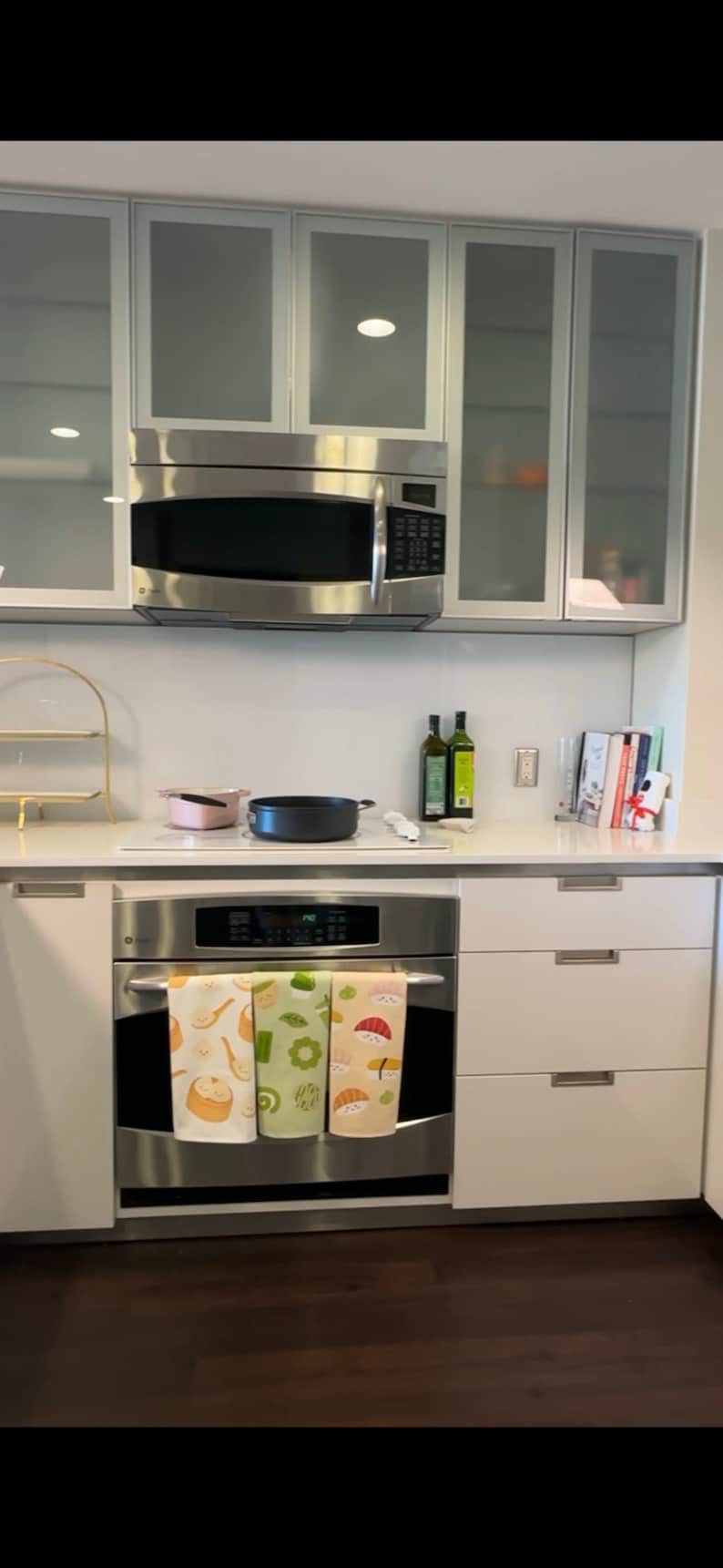 May include: A stainless steel oven with three kitchen towels hanging on the front. The towels have a sushi theme with images of sushi rolls, seaweed, and chopsticks. The oven is in a modern kitchen with white cabinets and a stainless steel microwave above it.