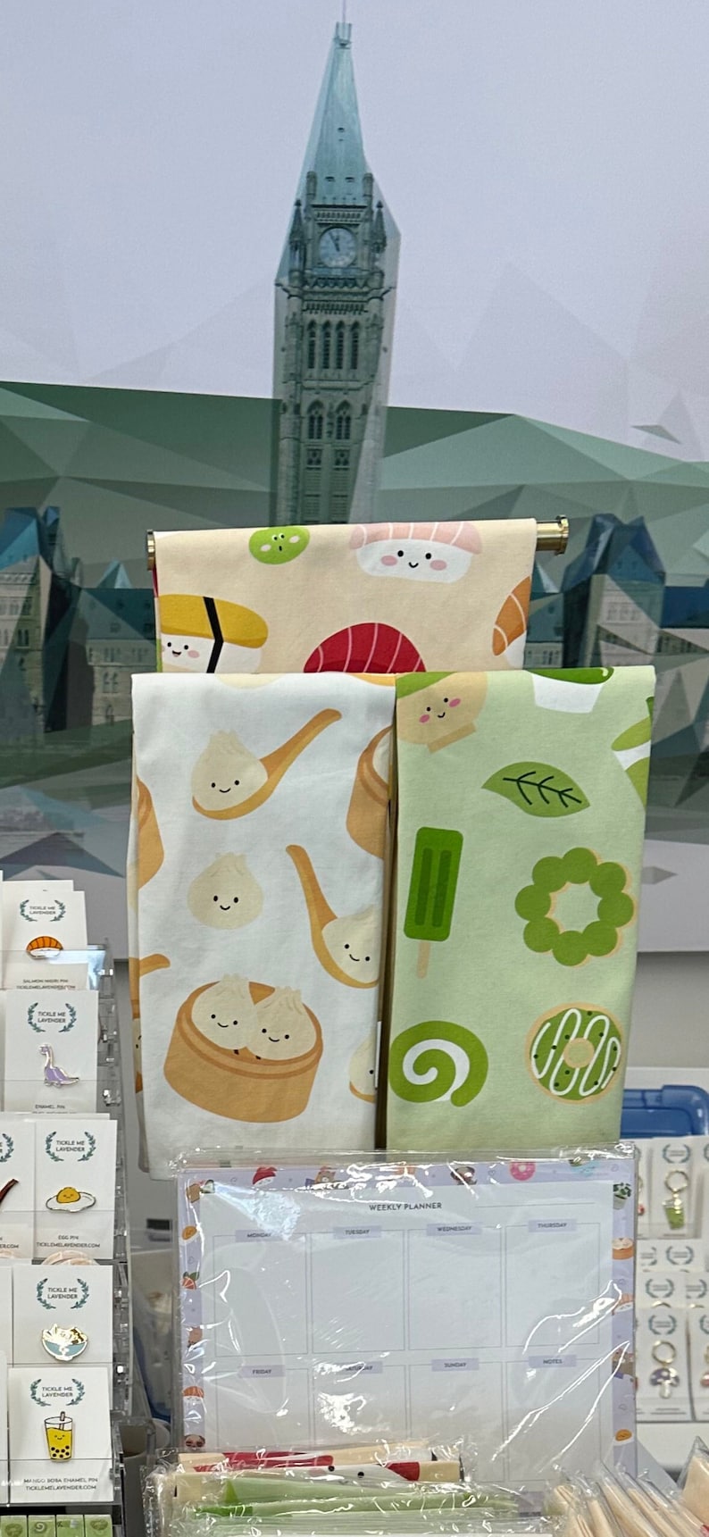 May include: Three kitchen towels with cute cartoon food designs. The towels feature sushi, dumplings, and green tea ice cream. A weekly planner with a white background and a colorful border is in the foreground.