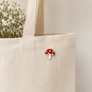 May include: A red and white mushroom enamel pin with a white border is attached to a beige canvas tote bag. The mushroom pin has a red cap with white spots and a white stem. The tote bag is filled with white flowers.