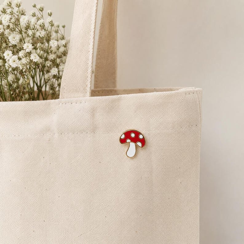 May include: A red and white mushroom enamel pin with a white border is attached to a beige canvas tote bag. The mushroom pin has a red cap with white spots and a white stem. The tote bag is filled with white flowers.