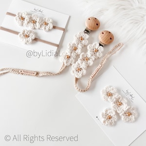 May include: Set of three crocheted flower headbands and a crocheted flower pacifier clip with wooden beads and clips. The set is in cream and gold colors. The text "byLidiAL" is on the packaging.