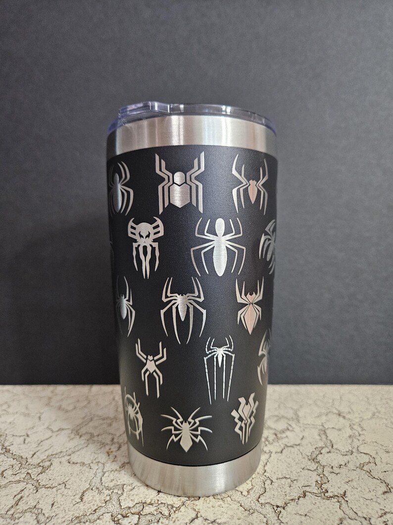 Puede incluir: Un vaso de acero inoxidable negro con varios dise&ntilde;os grabados en el lateral, con tem&aacute;tica de Spider-Man.