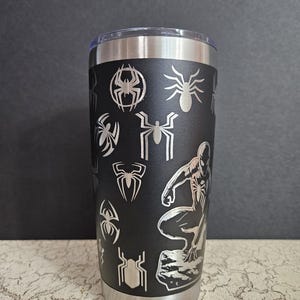 Puede incluir: Un vaso de acero inoxidable negro con un dise&ntilde;o de Spider-Man plateado. El vaso est&aacute; cubierto con un patr&oacute;n de ara&ntilde;as y siluetas de Spider-Man.