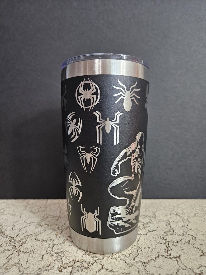 Puede incluir: Un vaso de acero inoxidable negro con un dise&ntilde;o de Spider-Man plateado. El vaso est&aacute; cubierto con un patr&oacute;n de ara&ntilde;as y siluetas de Spider-Man.