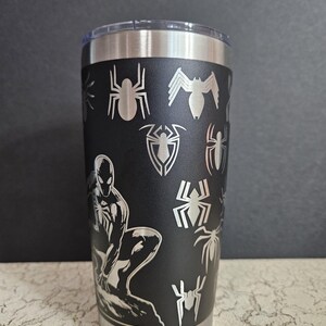 Puede incluir: Un vaso de acero inoxidable negro con tapa plateada. El vaso tiene un dise&ntilde;o negro y plateado con un personaje de Spider-Man y varias siluetas de ara&ntilde;as.
