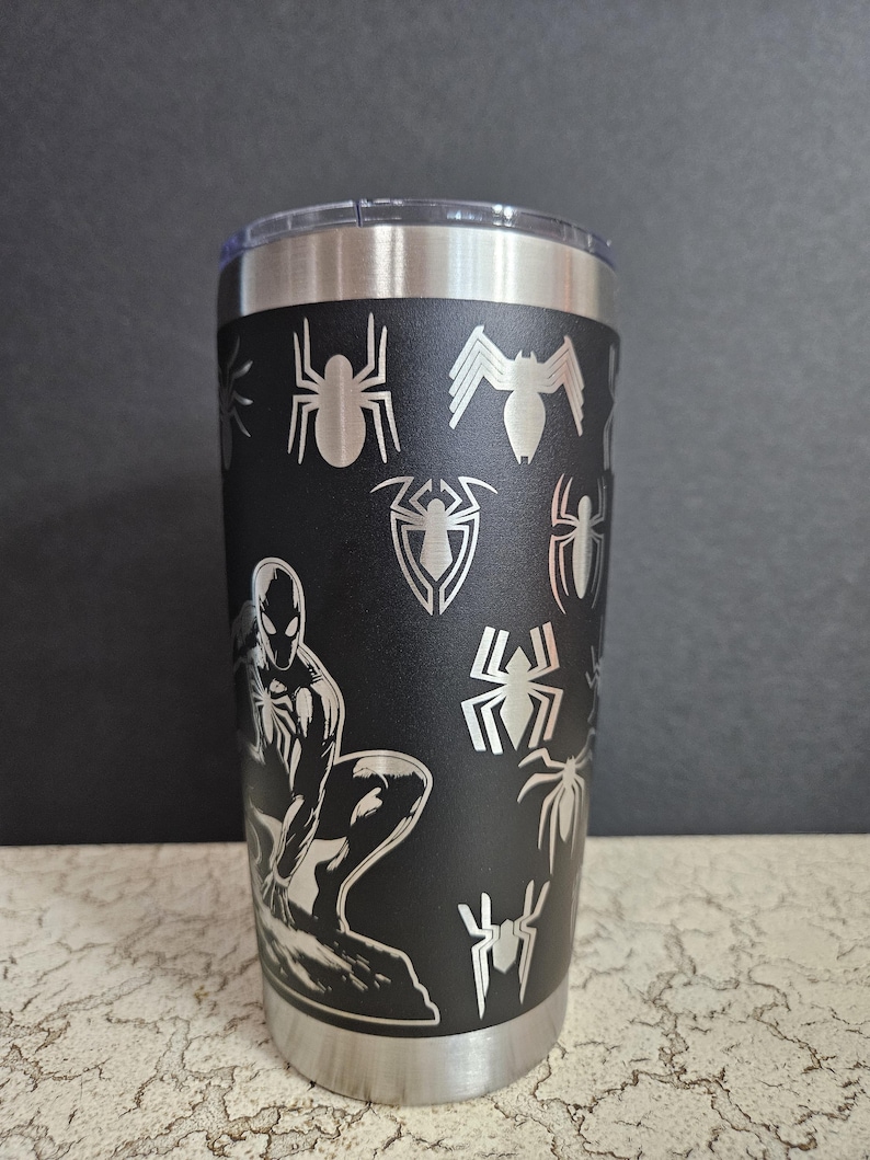 Puede incluir: Un vaso de acero inoxidable negro con tapa plateada. El vaso tiene un dise&ntilde;o negro y plateado con un personaje de Spider-Man y varias siluetas de ara&ntilde;as.