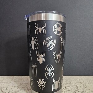 Puede incluir: Un vaso de acero inoxidable negro con una tapa plateada. El vaso est&aacute; cubierto con un patr&oacute;n de siluetas de ara&ntilde;as plateadas.