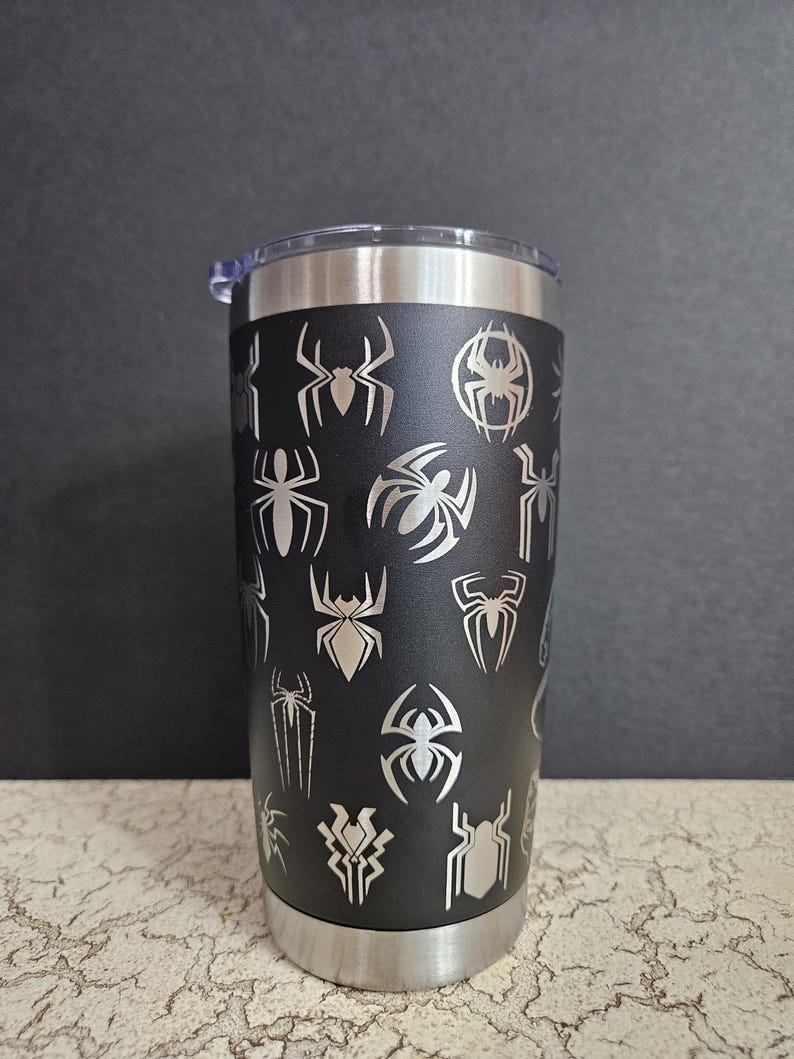 Puede incluir: Un vaso de acero inoxidable negro con una tapa plateada. El vaso est&aacute; cubierto con un patr&oacute;n de siluetas de ara&ntilde;as plateadas.