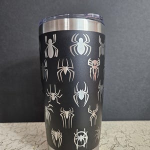 Puede incluir: Un vaso de acero inoxidable negro con un dise&ntilde;o de telara&ntilde;a. El vaso tiene una tapa plateada y una base plateada.