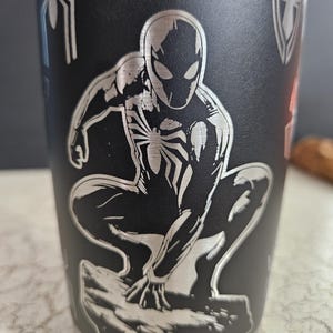 Puede incluir: Un vaso de acero inoxidable negro con una imagen grabada en plata de Spider-Man en posici&oacute;n de cuclillas. El vaso tiene una tapa plateada y un fondo plateado.